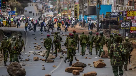 Getty Images A group of military police officers with guns run towards a groups of protesters. The road in between them is littered with rocks